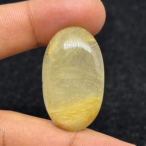以下が含まれることがあります： 金色の針状のインクルージョンが入った、楕円形の半透明の宝石。磨かれた石は温かみのある黄色で、暗い背景を背景に指で持たれています。宝石の長さは約2.5cmです。