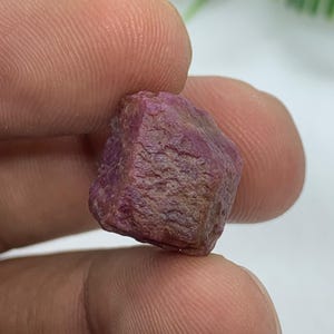 May include: A raw, uncut ruby gemstone with a deep magenta and reddish-purple hue. The stone features a naturally textured, irregular surface with visible crystalline facets and opaque sections, showcasing its unpolished and organic state.