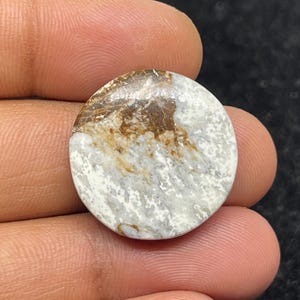 Peut inclure: Une pierre précieuse ronde et polie, présentant un motif marbré. La pierre combine des couleurs blanches, marron clair et beige. La pierre est tenue dans une main, mettant en valeur sa taille et sa texture. Elle est probablement destinée à la bijouterie ou à la décoration.