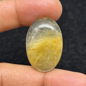 以下が含まれることがあります： 透明なベースと金色の針状のインクルージョンを持つ楕円形の宝石。暗い背景に対して、2本の指の間に石が保持されています。宝石のインクルージョンは、きらめく効果を生み出します。