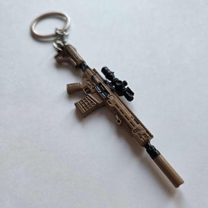 May include: A miniature rifle keychain in shades of brown and black. The keychain features a detailed replica of a firearm, complete with a scope and magazine. The metal ring is attached to the top of the rifle.