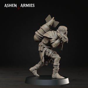 May include: A grey unpainted miniature figure from Ashen Armies, depicting a character carrying a large bomb on their back with rope bindings. The figure wears tattered clothing, leg wraps, and features cross symbols on their belt and gear.