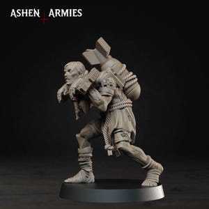 May include: A grey miniature figurine of a hunched figure carrying a large bomb on their back, secured with thick rope. The figure wears tattered clothing and wraps around their legs. The text ASHEN ARMIES is visible in the top left corner.