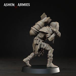 May include: A grey miniature figurine of a hooded figure in a crouching pose, carrying a large bomb strapped to their back with rope. The figure wears tattered clothing with cross symbols and leg wrappings. The text ASHEN ARMIES is visible.
