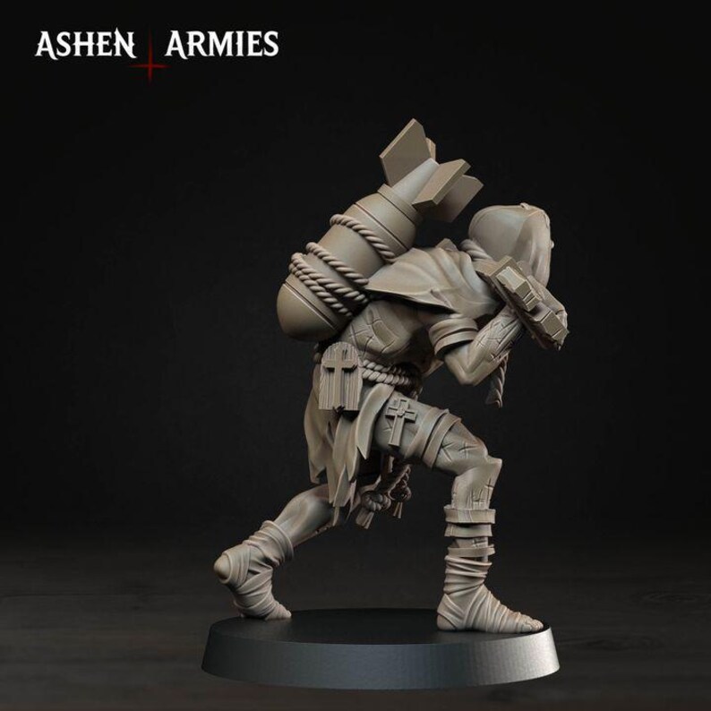 May include: A grey miniature figurine of a hooded figure in a crouching pose, carrying a large bomb strapped to their back with rope. The figure wears tattered clothing with cross symbols and leg wrappings. The text ASHEN ARMIES is visible.