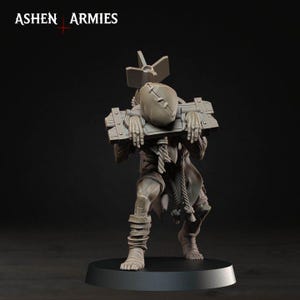 May include: A grey miniature figure of a humanoid creature with a sack-like head, wearing a wooden pillory around its neck and wrists. A large bomb-shaped object is strapped to its back. The text ASHEN ARMIES is visible in the upper left corner.