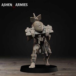 May include: A grey miniature figurine of a humanoid figure wearing a sack-like head covering and a wooden pillory, with a bomb-shaped object attached to the back. The text ASHEN ARMIES is visible in the upper left corner of the image.