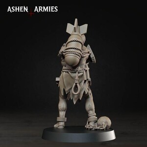 May include: A grey unpainted miniature figure standing on a circular base, featuring the text ASHEN ARMIES at the top. The figure carries a large bomb strapped to its back with rope, wears tattered clothing, and has a ball and chain attached to one leg.