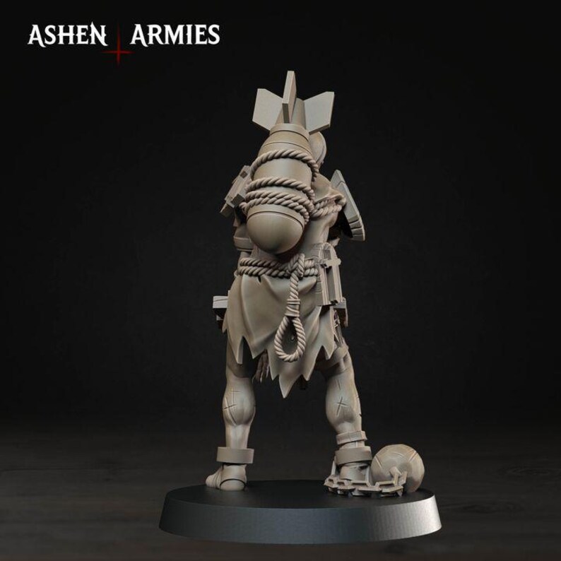 May include: A grey unpainted miniature figure standing on a circular base, featuring the text ASHEN ARMIES at the top. The figure carries a large bomb strapped to its back with rope, wears tattered clothing, and has a ball and chain attached to one leg.