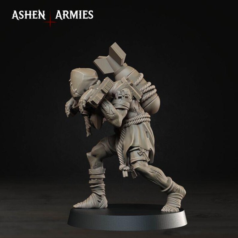 May include: A grey unpainted miniature figure from Ashen Armies, depicting a hunched character carrying a large bomb on their back secured with rope. The figure wears tattered clothing, leg wraps, and a mask, standing on a circular black base.