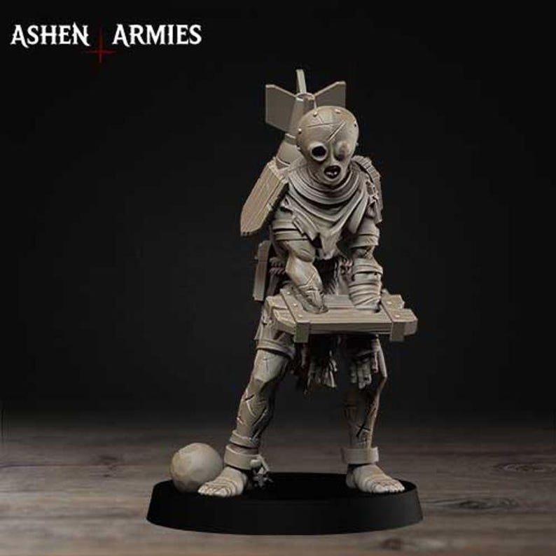May include: A light gray miniature figurine labeled ASHEN ARMIES, depicting a humanoid figure with a mask, wearing tattered clothing, carrying a large bomb on its back, and holding a wooden crate, standing on a circular black base with a ball.