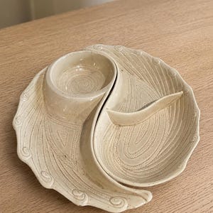 May include: A cream-colored ceramic serving dish with a glossy finish and speckled texture. The dish features a divided design with a circular bowl on one side and a curved, open section on the other, both decorated with intricate, embossed swirl patterns.