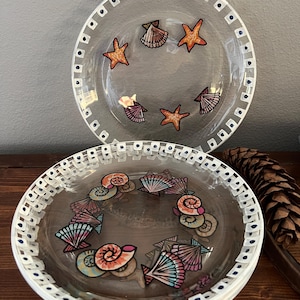 May include: Clear glass plates with seashell and starfish designs. The plates have a white border with blue dots. The seashell designs include orange, pink, and teal colors. The plates are stacked on top of each other.
