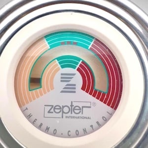 May include: Close-up of a Zepter Thermo Control device. The device has a white face with a circular design featuring teal, red, and tan arcs. The text "Zepter International" and "Thermo.Control" are printed on the face. The device is surrounded by a silver metal ring.