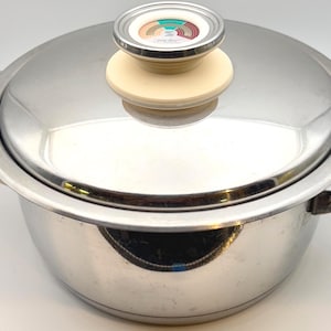 May include: Stainless steel cooking pot with lid. The pot has two handles and a lid with a cream-colored knob and a temperature gauge. The pot is reflective and sits on a white surface.