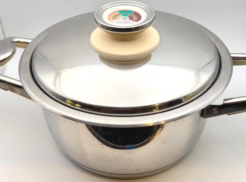 May include: Stainless steel cooking pot with lid. The pot has two handles and a lid with a cream-colored knob and a temperature gauge. The pot is reflective and sits on a white surface.