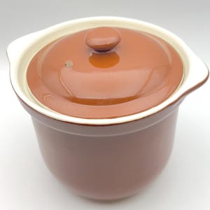 May include: A brown ceramic cooking pot with a matching lid. The pot has a rounded shape with a white rim and handles. The lid has a small knob on top. The pot is designed for cooking and serving.