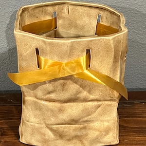 May include: A beige ceramic vase designed to resemble a crumpled paper bag with a textured, matte finish. The vase features vertical slits near the top rim, through which a bright yellow satin ribbon is threaded and tied in a bow at the front.
