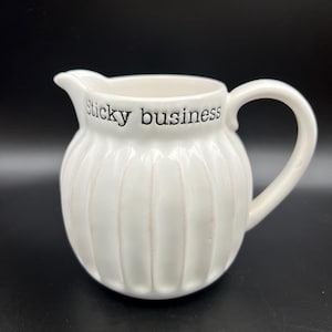 May include: White ceramic pitcher with a rounded body and a handle. The pitcher has vertical ribbed detailing and the words "sticky business" in black lettering. The spout is small and curved.