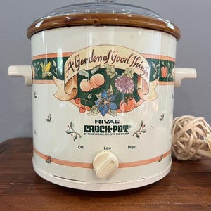 May include: A cream-colored Rival Crock-Pot stoneware slow cooker with a glass lid. The exterior features a decorative band with the text A Garden of Good Things and illustrations of fruits and vegetables. It has a single control knob for Off, Low, and High settings.