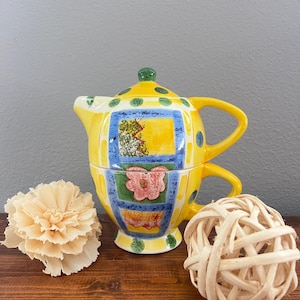 May include: A yellow ceramic tea-for-one set featuring a teapot stacked on a teacup. The set is decorated with blue grid patterns, green polka dots, and colorful floral motifs. The teapot has a green lid knob and a curved handle.
