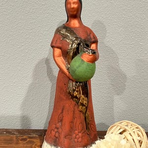 May include: A terracotta-colored ceramic figurine of a person with black hair wearing a long dress with a floral pattern at the hem. The figure has a metallic bronze-colored sash draped over the shoulder and holds a round, textured green vessel.