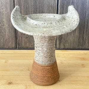 May include: A speckled ceramic bird bath with a wide, shallow basin and two curved handles. The base is a light beige color, transitioning to a brown base. The bird bath is a decorative item for the garden.