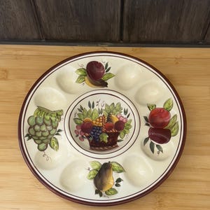May include: A round, off-white ceramic serving dish with a maroon rim. The dish features a central fruit basket design and individual fruit illustrations, including grapes, apples, and pears, around the perimeter. It has ten recessed sections for serving.