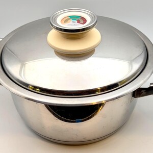 May include: Stainless steel cooking pot with lid. The pot has a shiny, reflective surface and two side handles. The lid features a cream-colored handle with a decorative top.