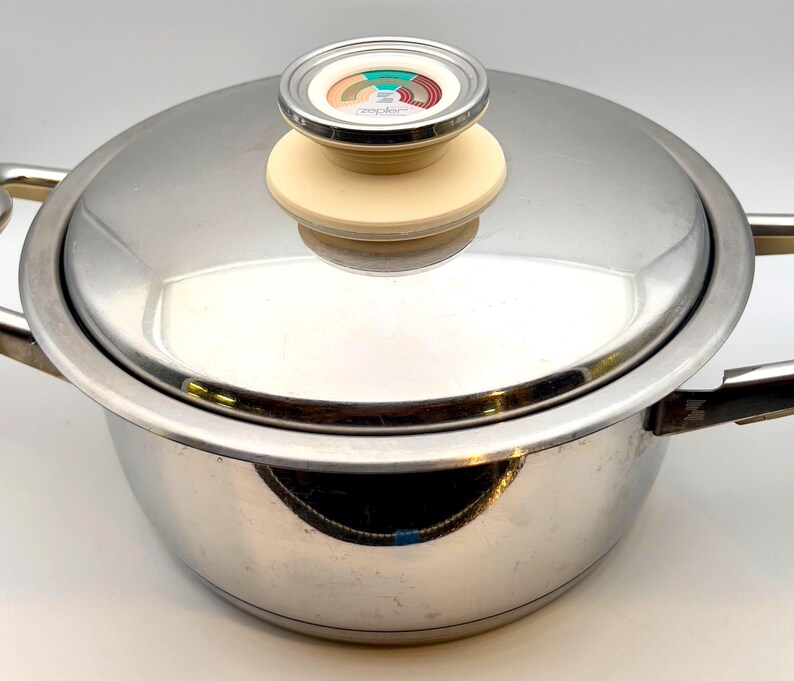 May include: Stainless steel cooking pot with lid. The pot has a shiny, reflective surface and two side handles. The lid features a cream-colored handle with a decorative top.