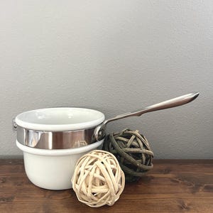 May include: A white saucepan with a stainless steel handle and rim. Two decorative spheres, one light-colored and one dark green, sit beside the pan. The objects are on a dark brown wooden surface against a gray wall.