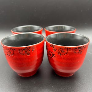 May include: Four red ceramic bowls with black interiors. The bowls have a textured finish and a black floral pattern near the rim. The bowls are arranged in a square formation against a dark background.