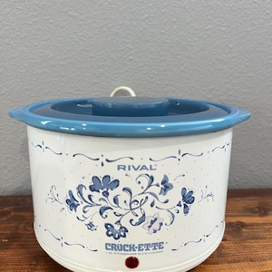 May include: A white Rival Crock-Ette 1 qt. stoneware slow cooker with a blue lid and a blue floral pattern printed on the front. The appliance features a small red indicator light near the base and a power cord extending from the back.