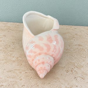 May include: A ceramic seashell-shaped planter or decorative vessel featuring a glossy finish with a white base and soft pink mottled patterns. The object has a spiral-shaped exterior and a smooth, hollow interior opening at the top.