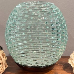 May include: A spherical lamp featuring a light blue-green glass exterior constructed from numerous small, rectangular glass pieces arranged in a brick-like pattern. The glass sphere rests upon a dark brown circular base, creating a textured surface.