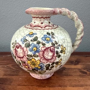 May include: A ceramic pitcher with a cream-colored crackle glaze finish, featuring a hand-painted floral pattern in shades of pink, blue, and yellow. The vessel has a rounded body, a flared rim, and a twisted handle with pink accents.