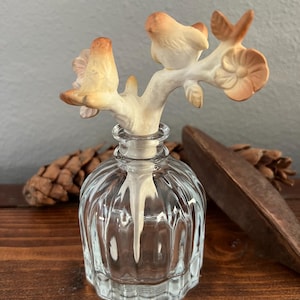 May include: A clear glass bottle with a decorative stopper featuring two small birds perched on a branch with flowers. The birds and branch are painted in shades of beige and brown. The bottle has a ribbed design.