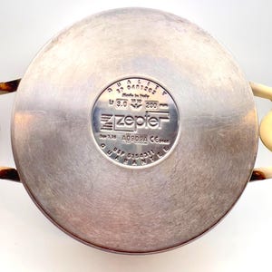 May include: A stainless steel Zepter pot base with cream-colored handles. The pot has a circular design with a central logo and text, including measurements of 200 mm. The pot is made in Italy.
