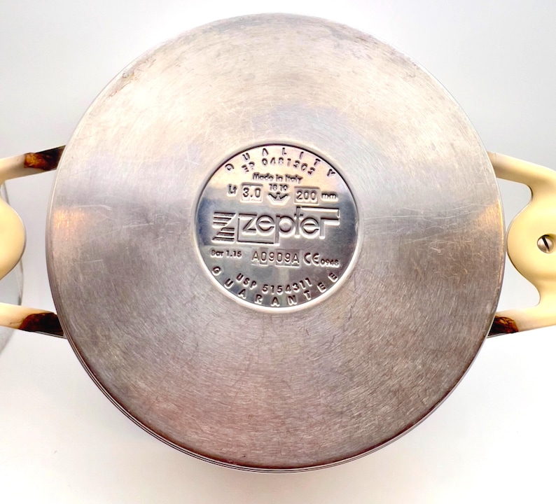 May include: A stainless steel Zepter pot base with cream-colored handles. The pot has a circular design with a central logo and text, including measurements of 200 mm. The pot is made in Italy.