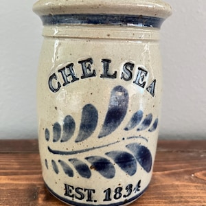 May include: A cylindrical stoneware crock with a speckled beige finish and a flared rim. The front features the embossed text CHELSEA in dark blue, a blue painted leaf motif, and the text EST. 1834 in dark blue at the base of the vessel.