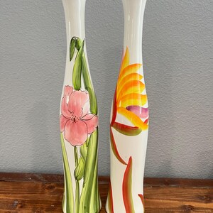 May include: Two tall, slender ceramic vases with a white glossy finish and a narrow neck. The left vase features a hand-painted pink iris with green stems and leaves. The right vase displays a vibrant orange and yellow bird of paradise flower.