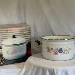 May include: A white enamel stockpot with black side handles and a floral pattern featuring pink and blue flowers. The pot is displayed next to its original cardboard box, which shows the text 34 CM MARMITE and Es Für Emaille-Kochgeschirr.