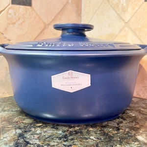 May include: A blue ceramic Emile Henry Dutch oven with a lid. The pot has two handles and a knob on the lid. A white label with text is on the front of the pot. The pot is sitting on a granite countertop.