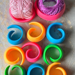 May include: An overhead shot features two spools of thread in pink and purple, each resting on a pink holder. Below, nine colorful stitch markers in blue, orange, pink, and green are arranged in a grid. These tools are for knitting and crochet projects.