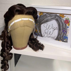 Może przedstawiać: Brązowa peruka z długimi, kręconymi lokami jest prezentowana na głowie manekina. Peruka ma koronkowy przód i jest zabezpieczona białą opaską. Widoczna jest oprawiona ilustracja z napisem "Wigs by Daniel".