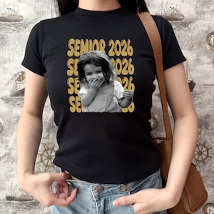 May include: Black t-shirt with a gold design featuring the text "SENIOR 2026" repeated vertically, and a black and white photo of a young child. The child is covering their mouth and smiling. The shirt is short-sleeved.