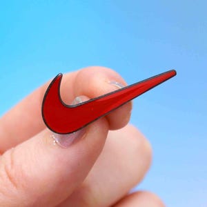 Peut inclure: Une broche en émail rouge en forme de logo Nike swoosh avec un fin contour noir. La broche présente une finition lisse et brillante ainsi qu'un design incurvé et effilé qui imite le symbole emblématique de la marque sportive en rouge.