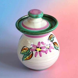 May include: A ceramic jar with a lid, featuring a white base with hand-painted pink flowers and green leaves. The lid has a green rim and a pink centre knob. The jar has a rounded body with subtle horizontal ridges and a glossy finish throughout.
