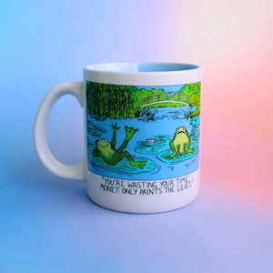 K&ouml;nnte beinhalten: Wei&szlig;e Keramik-Tasse mit einer Cartoon-Illustration von zwei Fr&ouml;schen in einem Teich mit Seerosen und einer Br&uuml;cke. Auf der Tasse steht der Text "YOU'RE WASTING YOUR TIME. MONET ONLY PAINTS THE LILIES."