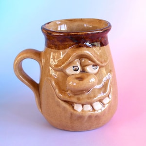 May include: A tan ceramic mug featuring a sculpted, expressive face with a wide, toothy grin, a prominent nose, and heavy eyelids. The mug has a glossy finish, a dark brown rim, and a curved handle on the left side, showcasing a hand-crafted design.